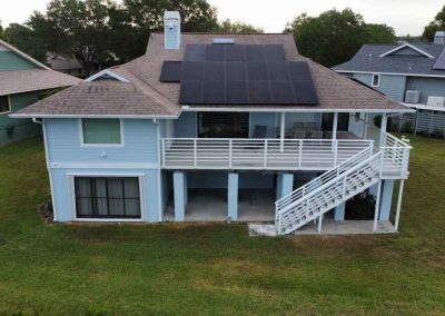 A coastal home with solar panels on the roof, surrounded by green grass and neighboring houses, seen from above.