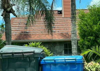 Residential roof with visible solar panel mounting hardware, surrounded by palm trees and trash bins in the foreground.