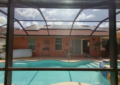 Solar panels installed on a home's roof, viewed through a screened enclosure, with a pool and patio area in the foreground.