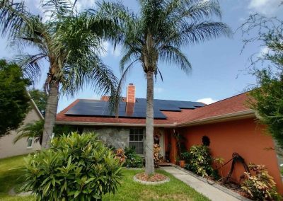 Solar panels installed on a home's red roof, surrounded by palm trees, green shrubs, and a landscaped front yard under clear skies.