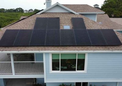 Aerial view of a coastal home with solar panels installed on the roof, overlooking greenery and a waterway.