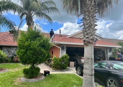 Workers on a residential roof installing solar panel mounts, with palm trees, a garage, and cars visible in the front yard.