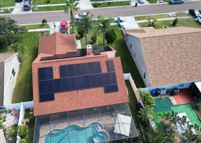 Solar panels covering a red-shingle residential roof, with a screened-in pool and surrounding neighborhood visible from above.